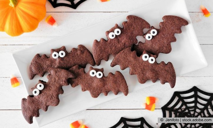 Süßes zu Halloween - Fledermaus-Plätzchen mit Honig auf einem Teller liegend. Im Hintergrund sind Spinnenweben und ein Kürbis zu sehen.