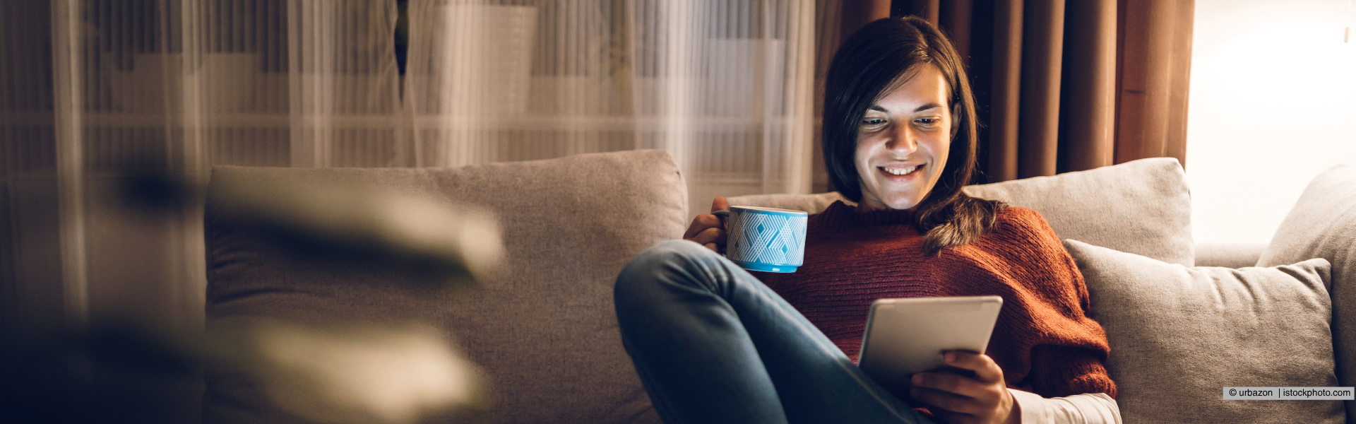 Eine Frau sitzt auf der Couch mit Tablett und einer Tasse in der Hand 