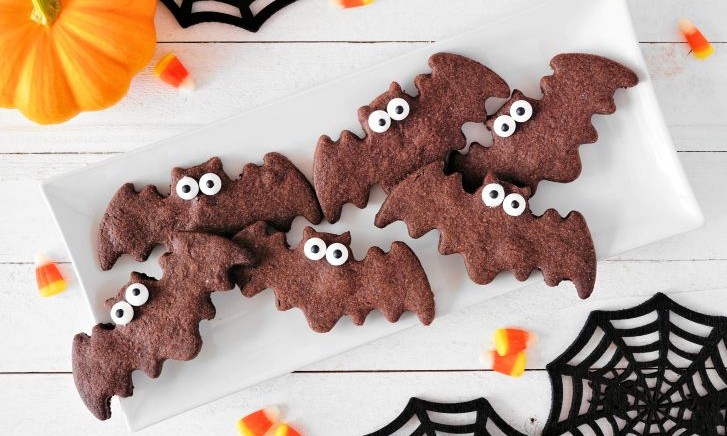 Süßes zu Halloween - Fledermaus-Plätzchen mit Honig auf einem Teller liegend. Im Hintergrund sind Spinnenweben und ein Kürbis zu sehen.