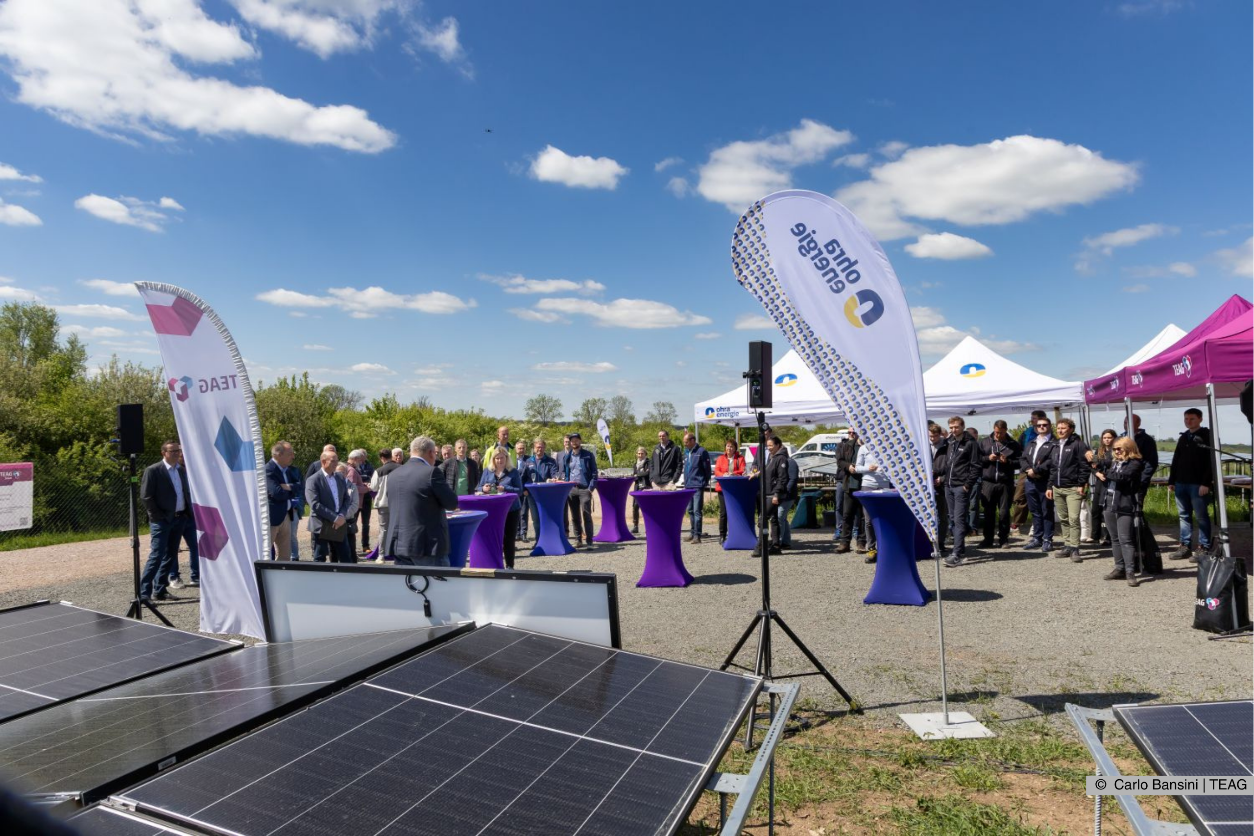 Die geladenen Gäste hören der Begrüßung zur Eröffnung des Solarparks aufmerksam zu. Die Beachflags der Ohra Energie und der TEAG wehen dabei im Wind. zur Eröffnung des Solarparks. Fotograf: Carlo Bansini/TEAG