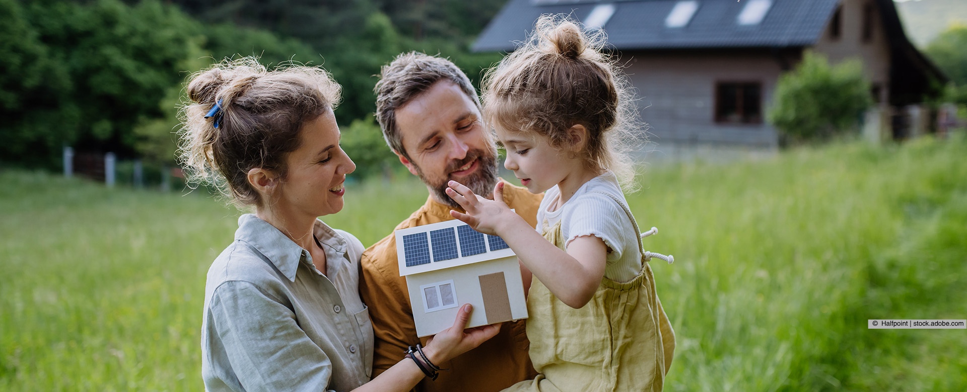 Familie steht auf einer Wiese vor einem Haus und hält ein Miniaturhaus in der Hand