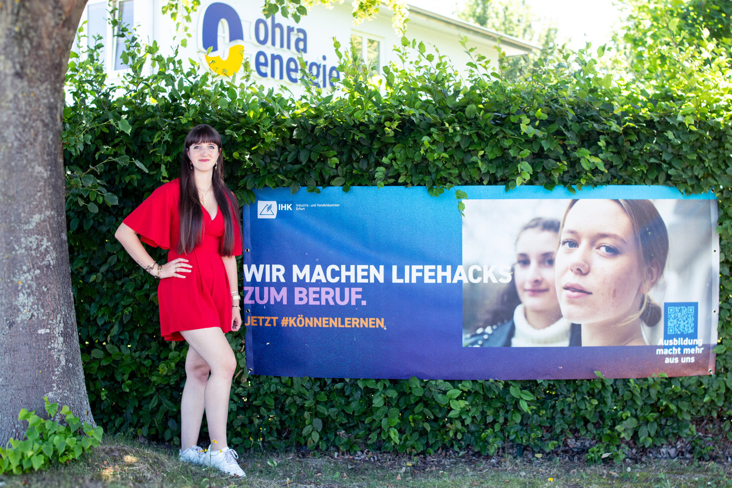 Elisa S. als Auszubildende vor der IHK-Banner "Wir machen Lifehacks zum Beruf", welcher an einem Busch hängt. Im Hintergrund das Firmengebäude der Ohra Energie zu sehen.