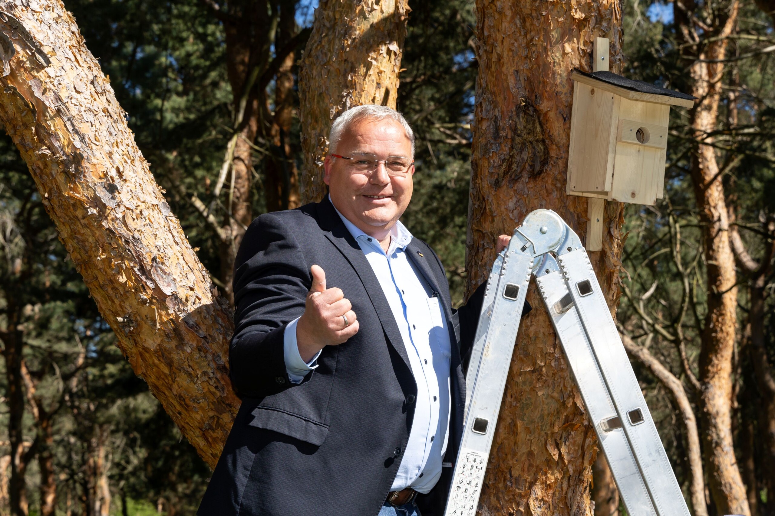 Herr Fischer steht auf einer leiter und blickt lächelns in die Kamera. Daneben ein Nistkasten am Baum. 
